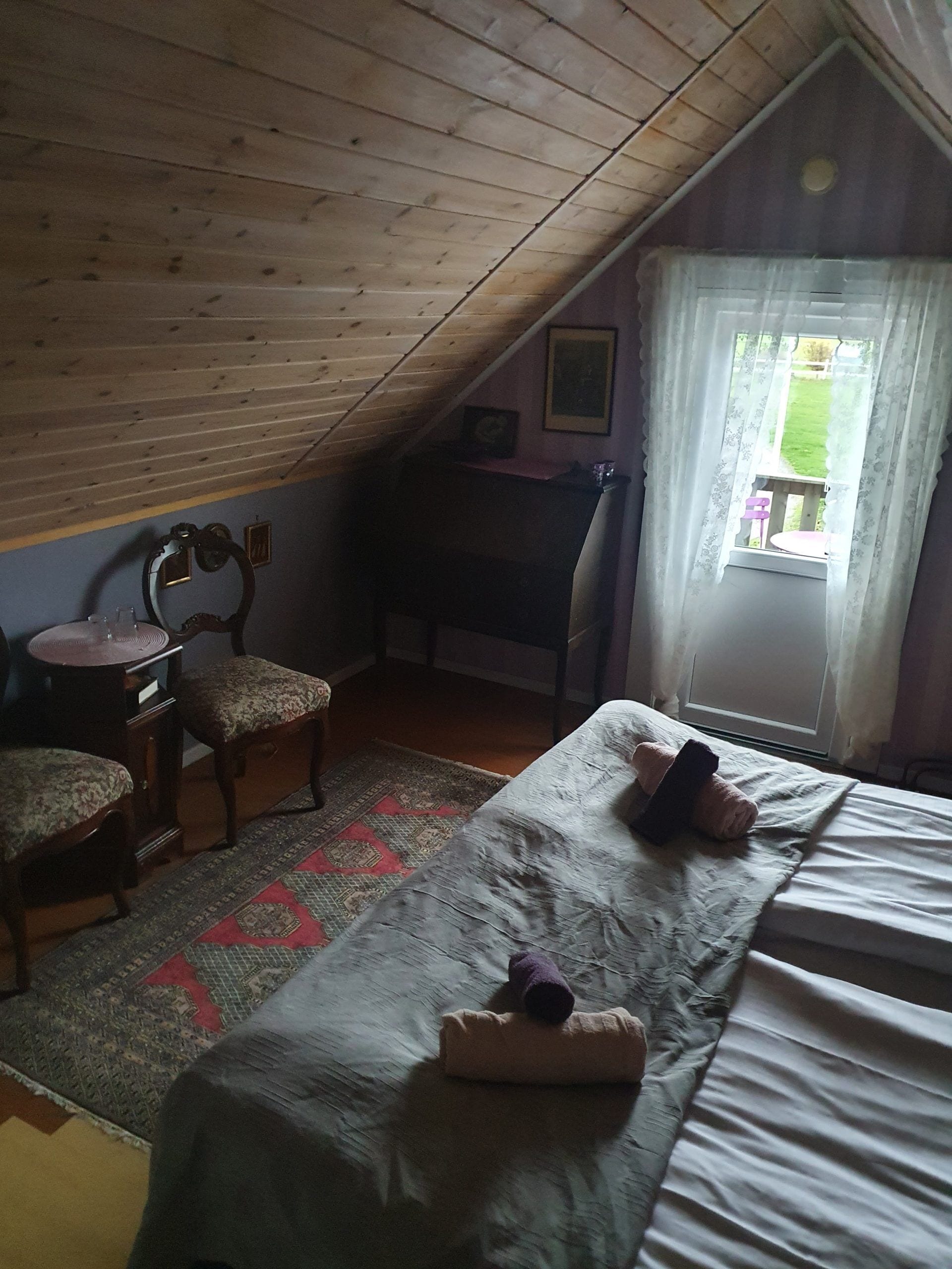Una camera da letto mansardata accogliente, con travi a vista, una scrivania in legno, una sedia, un letto con un asciugamano arrotolato e vista su un giardino verde attraverso una finestra.