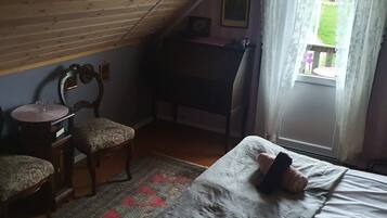 Una camera da letto mansardata accogliente, con travi a vista, una scrivania in legno, una sedia, un letto con un asciugamano arrotolato e vista su un giardino verde attraverso una finestra.