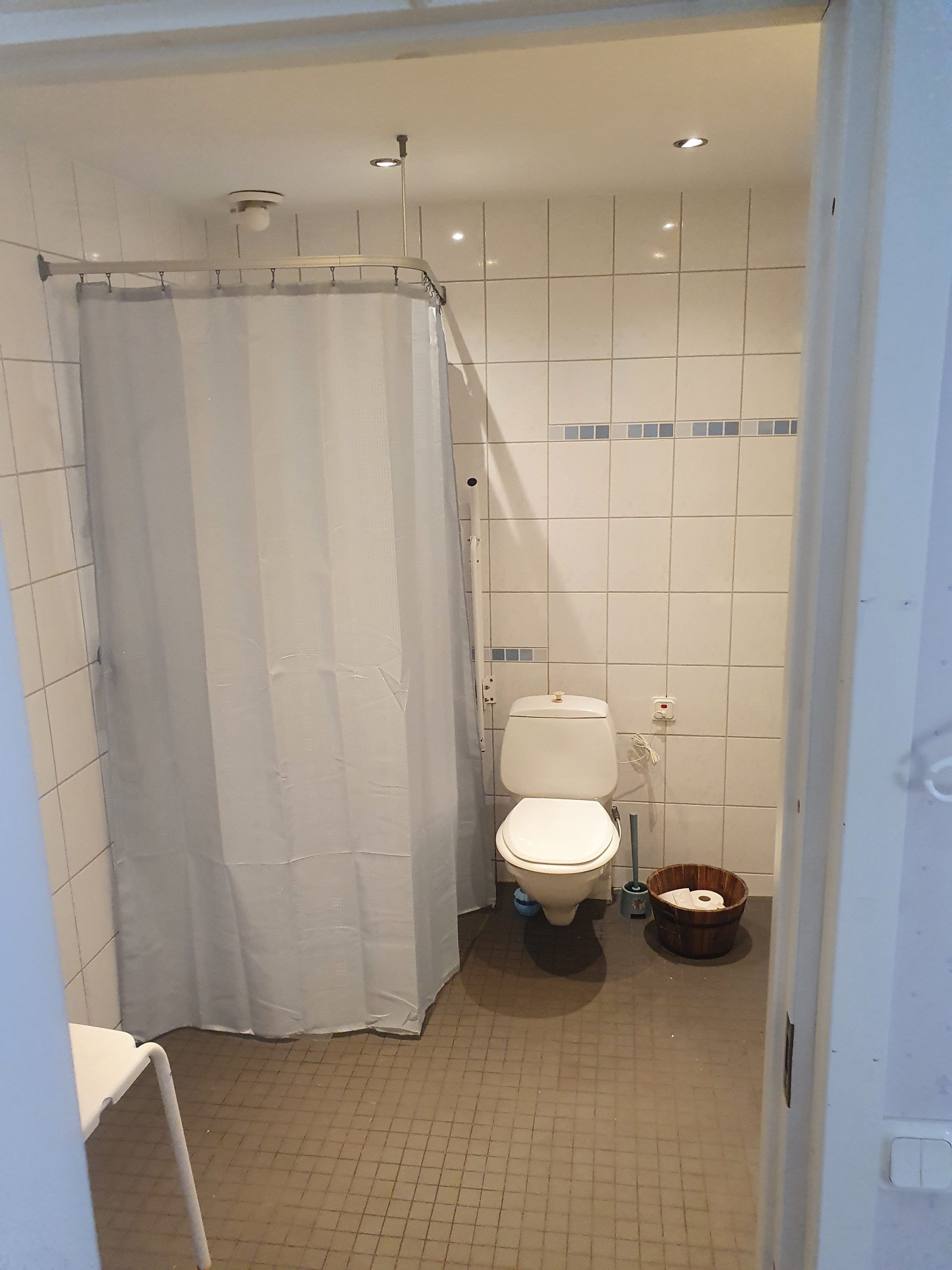 A bathroom with a toilet, a shower curtain, a small wooden bucket, and tiled walls.