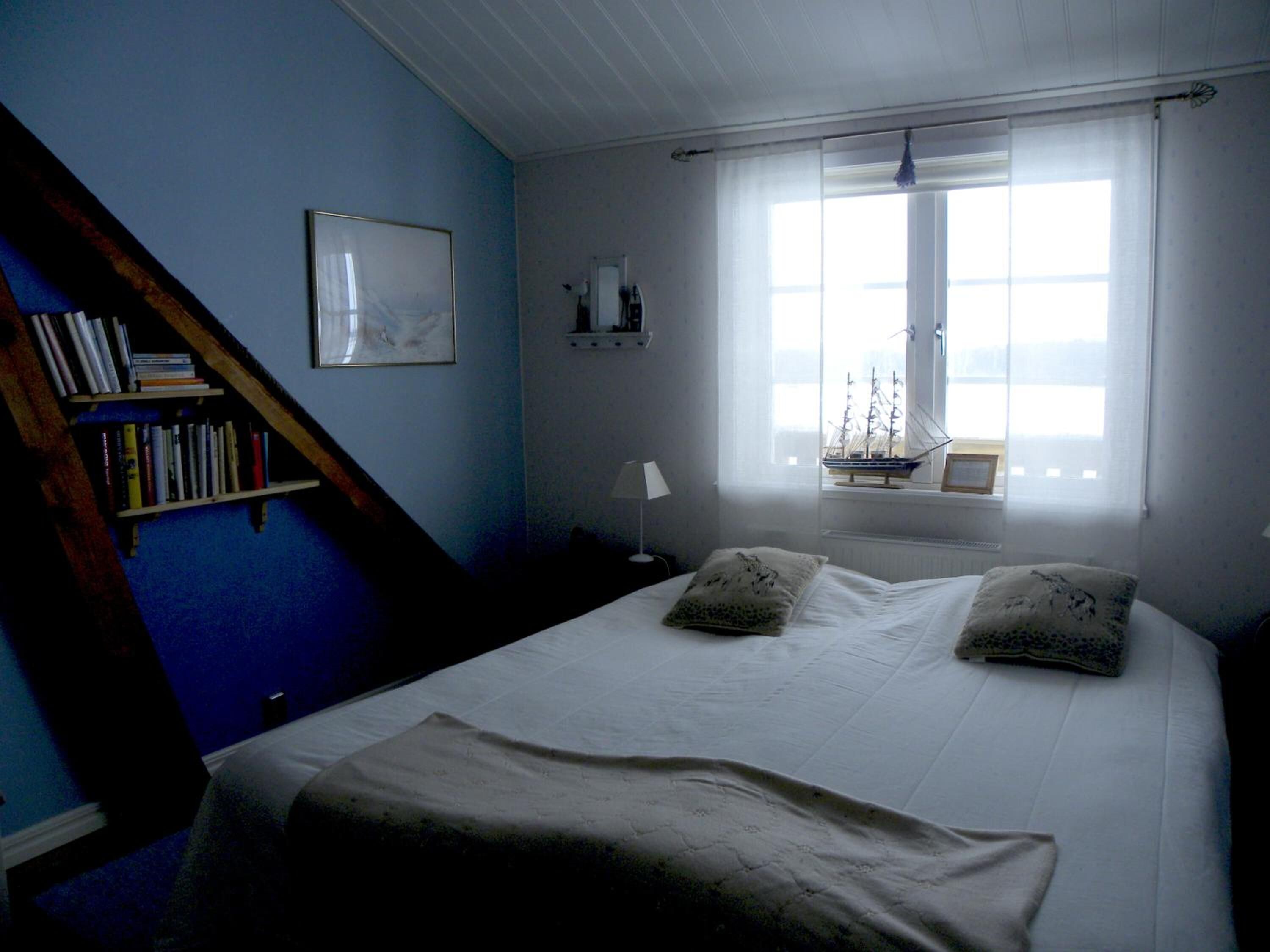 A bedroom with a bed, a bookshelf, a window with a ship model, and a painting on the wall.
