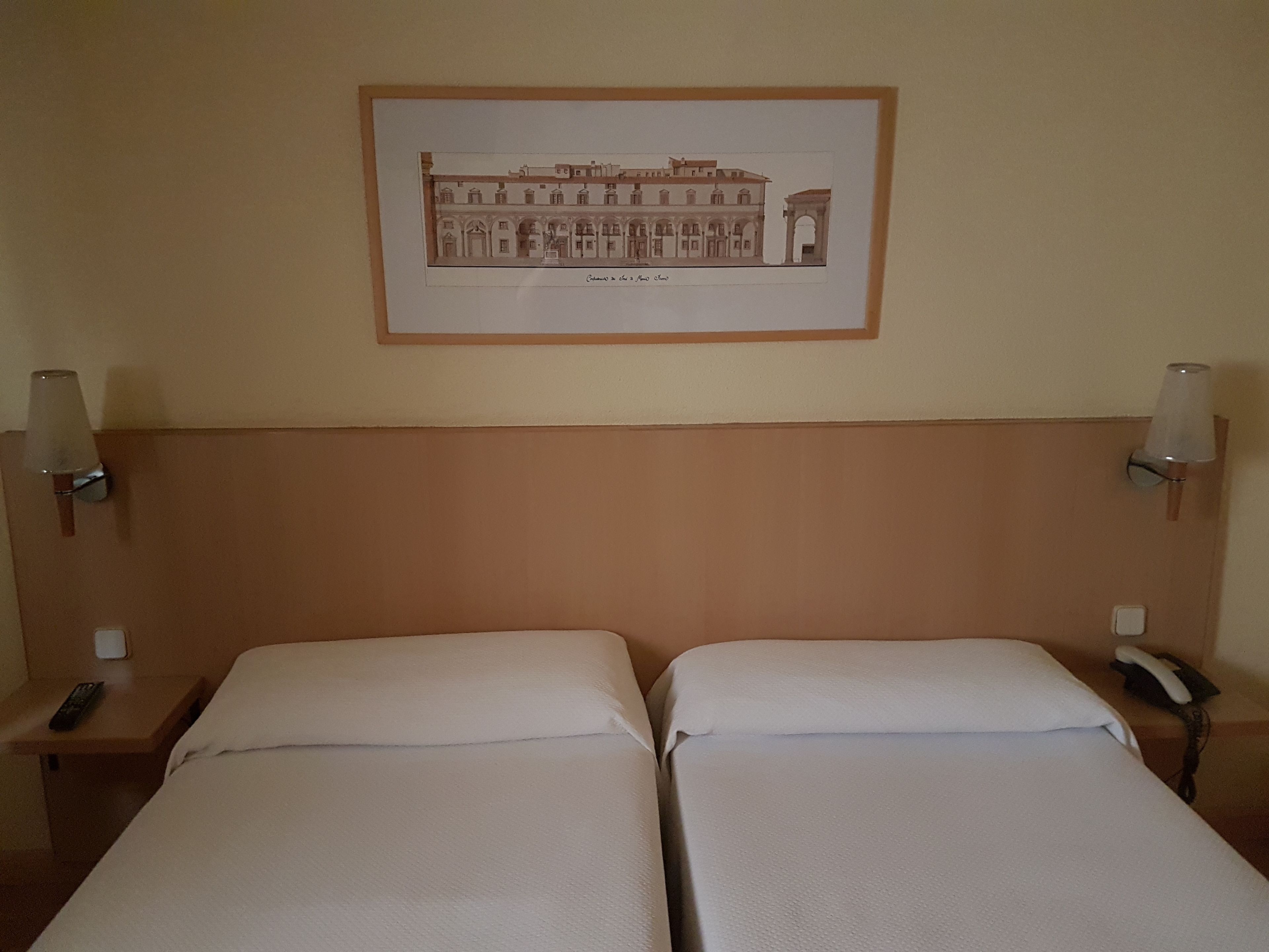 Habitación de hotel con dos camas individuales, un cuadro enmarcado de un edificio en la pared, lámparas de noche y un teléfono en la mesita.