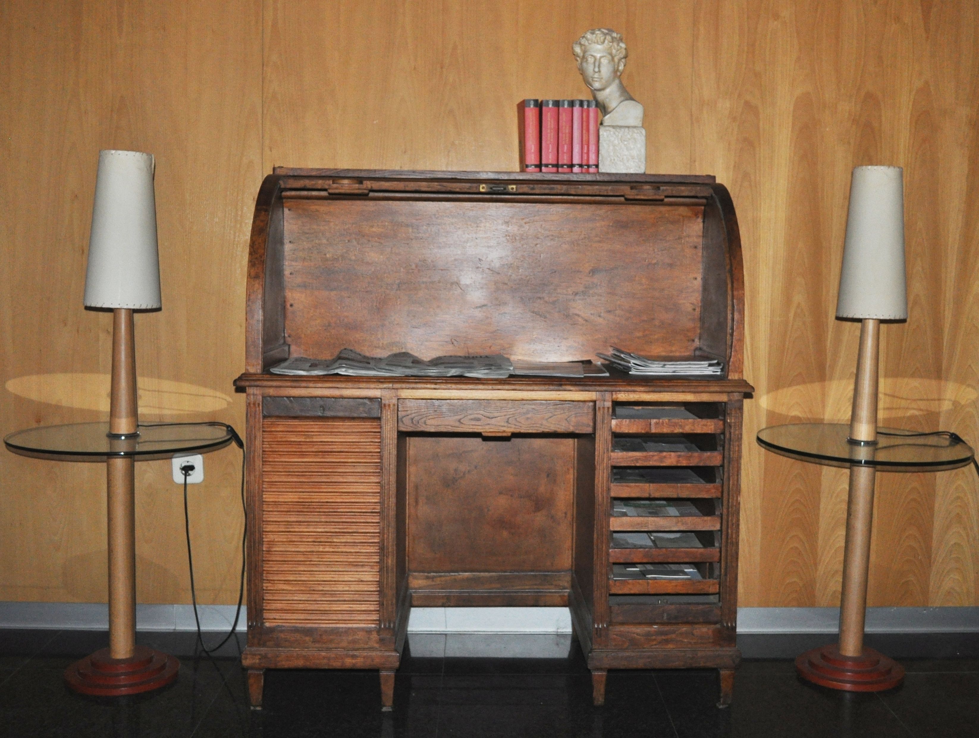 A wooden desk with a glass top, flanked by two lamps, and a bust sculpture on top.