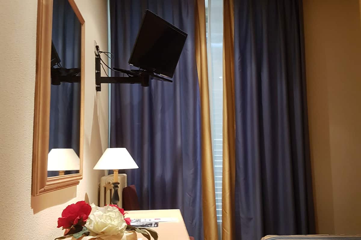 Une chambre d’hôtel avec un bureau en bois, un téléviseur à écran plat fixé au mur, une lampe, un miroir, un lit recouvert d’une courtepointe à rayures et une fenêtre avec des rideaux bleus.