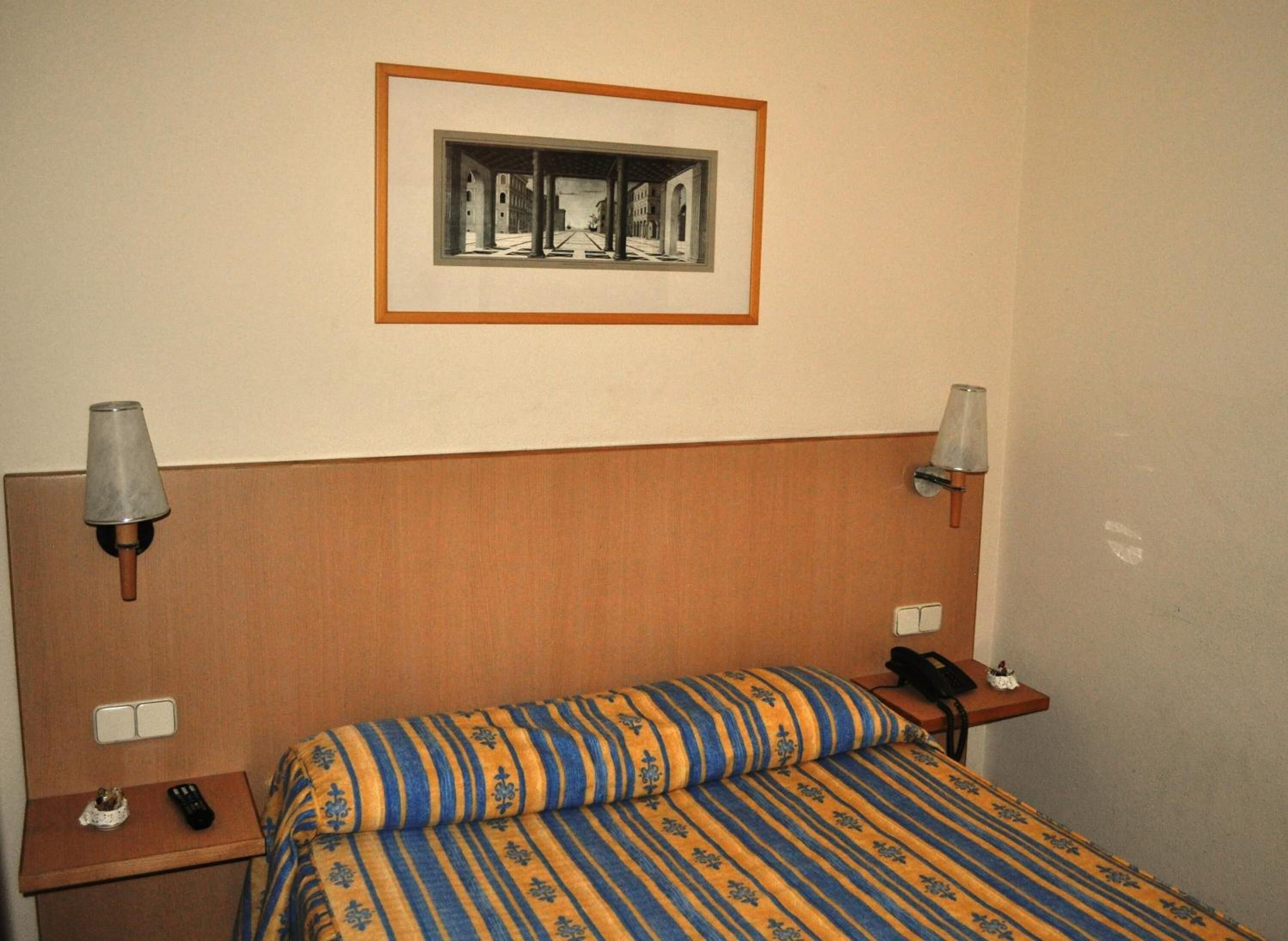 Habitación de hotel con cabecera de madera, cama con ropa de cama a rayas, dos mesitas de noche con lámparas, un cuadro enmarcado en la pared y un teléfono en una de las mesitas.