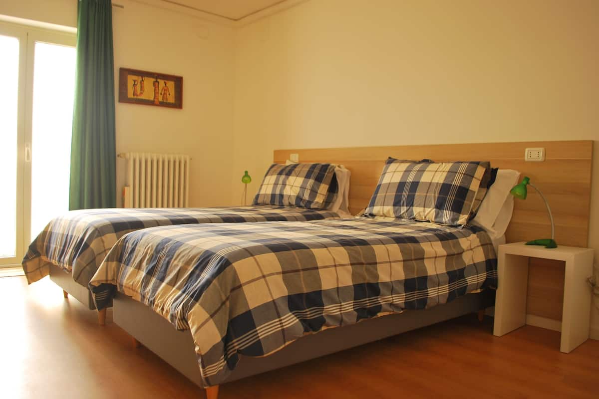 A double bed with a plaid bedspread, a bedside table with a lamp, and a radiator on the wall.