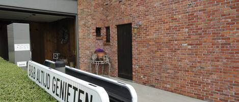 Un letrero de B&B Altijd Genieten, un edificio de ladrillo con una puerta de garaje, un ventanal pequeño y una mesa de metal con plantas.