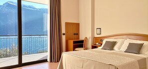 A hotel room with a large bed, a wooden nightstand, a desk, and a view of mountains and a body of water through a balcony. - Hotel Mercedes (Limone sul Garda)