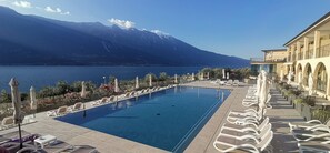 A swimming pool with lounge chairs and umbrellas, a mountain backdrop, and a building with balconies. - Hotel Mercedes (Limone sul Garda)