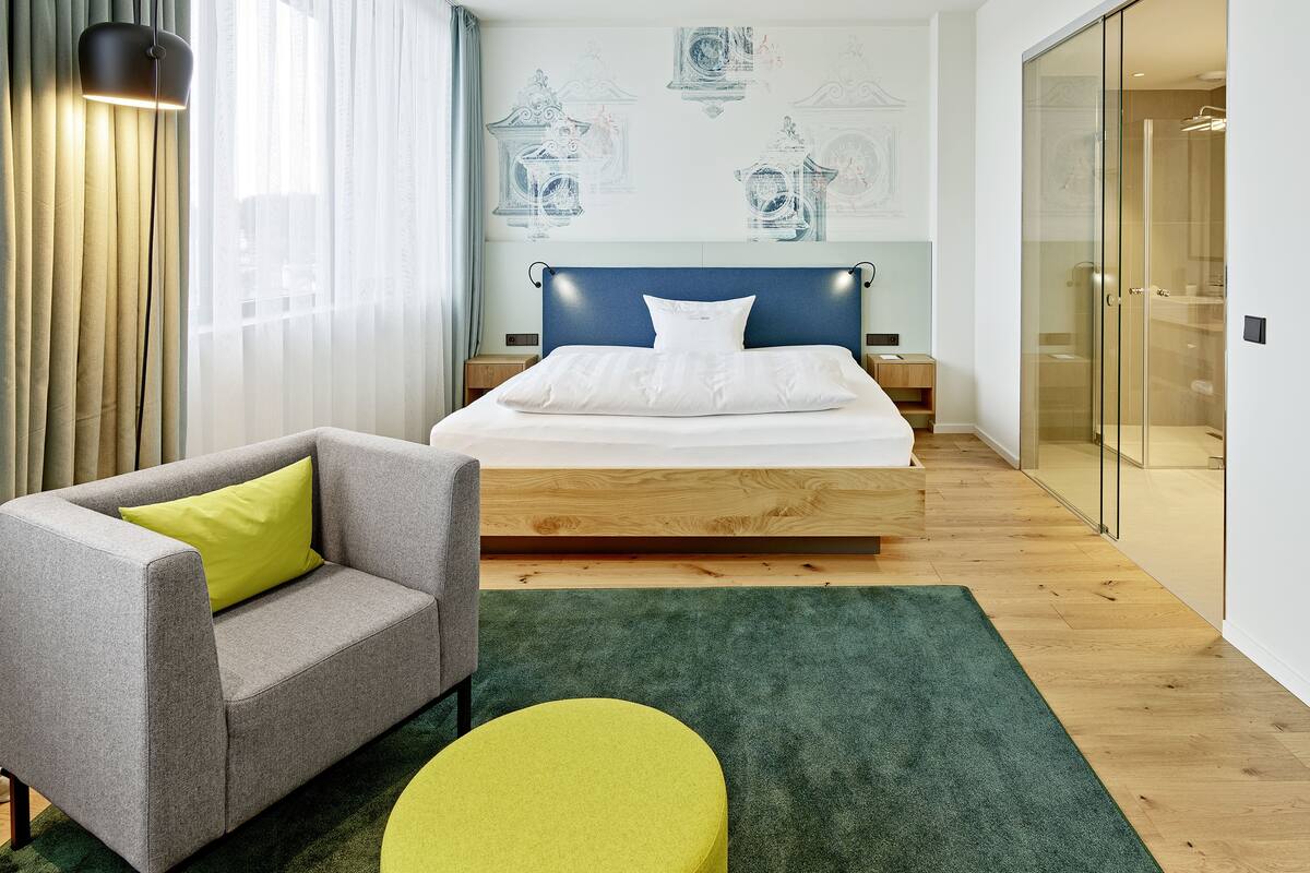 Una habitación de hotel moderna con una cama, un sofá, una alfombra verde y un box de ducha de vidrio.