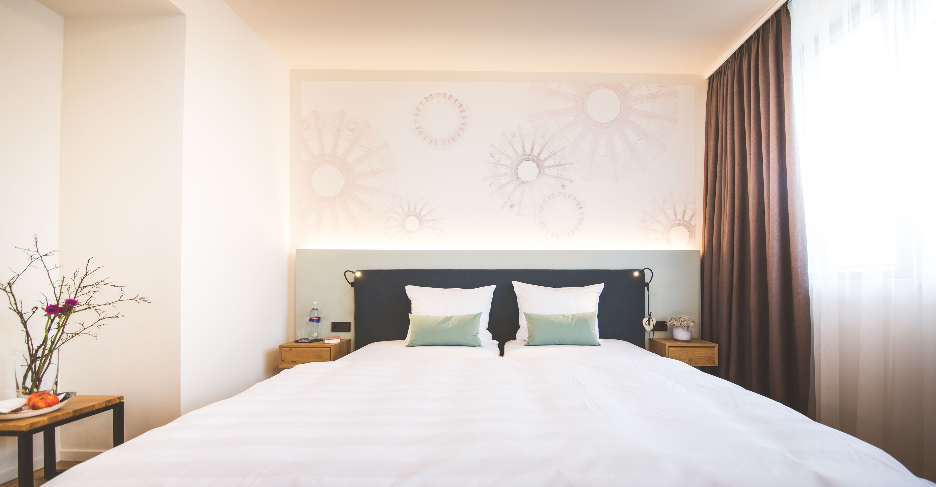 A hotel room with a large bed, two bedside tables, a wall-mounted light fixture, and a window with curtains.
