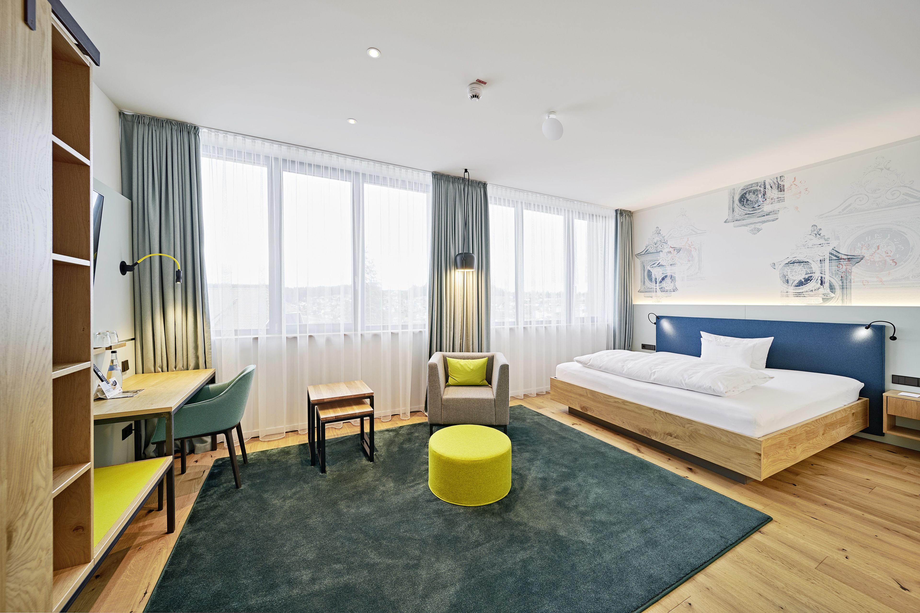 A modern hotel room with a wooden bed, a desk, a chair, and a green ottoman.