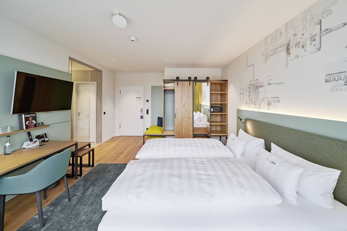 Una habitación de hotel moderna con una cama grande, un escritorio y un televisor. Se aprecia un armario con espejo y una estantería con una silla amarilla.