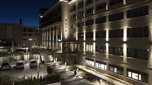 Ein Hotel mit modernem Design, das eine zentrale Treppe zum Eingang, einen Parkplatz mit mehreren Autos und eine gut beleuchtete Fassade zeigt.