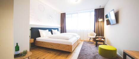 A modern hotel room with a wooden floor, a bed with white linens, a small round table with a bottle and glass, a yellow ottoman, a TV on the wall, and a window with curtains.