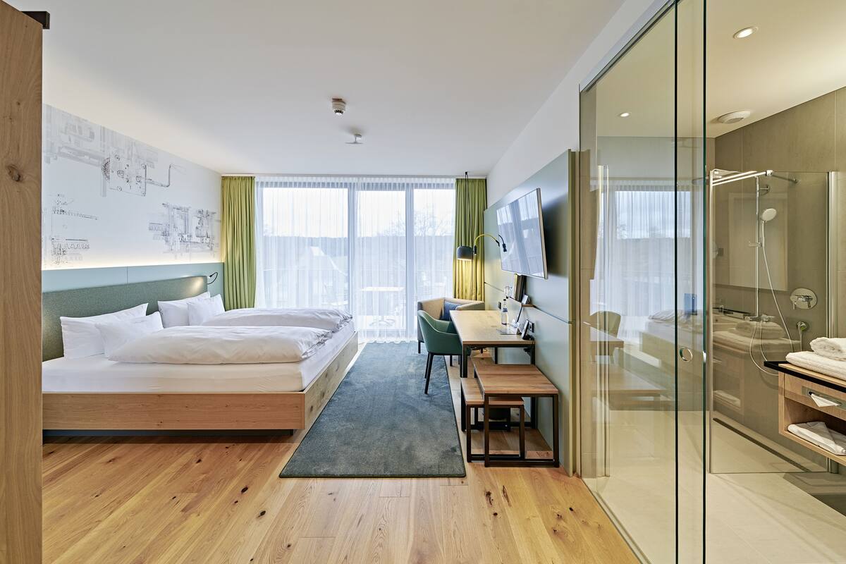 Una habitación de hotel moderna con cama, escritorio, silla y zona de ducha con paredes de vidrio.