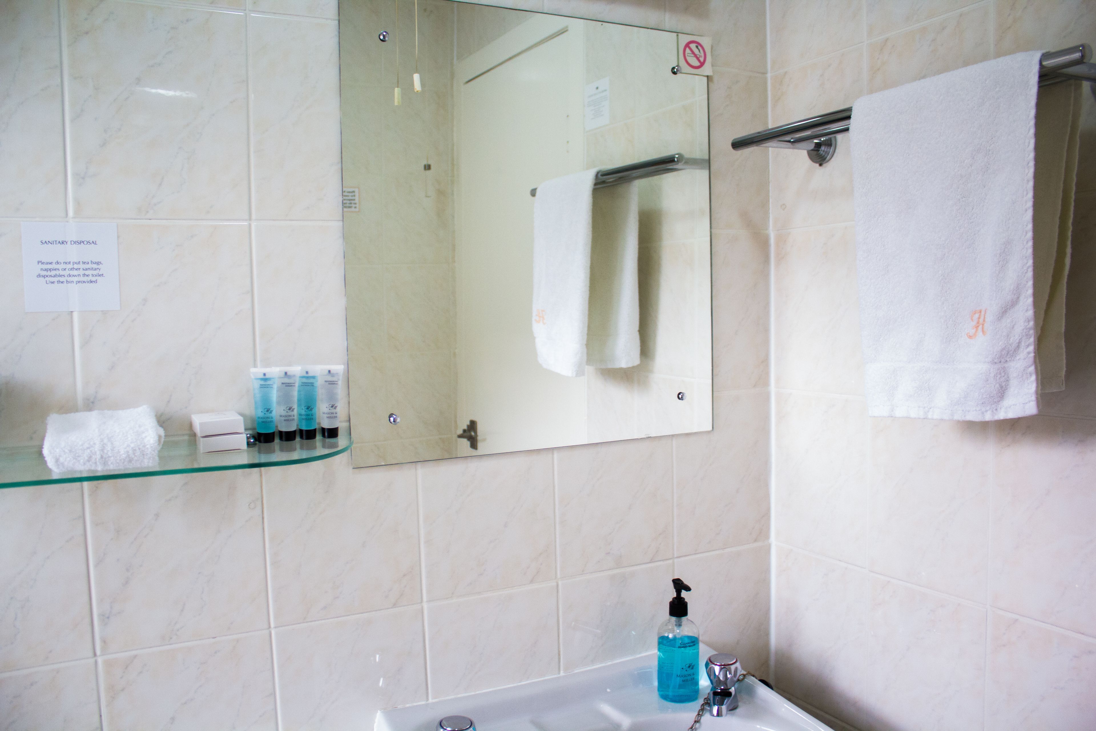 Un baño con un estante de vidrio que sostiene artículos de aseo, un lavamanos con dispensador de jabón y un perchero con una toalla blanca.