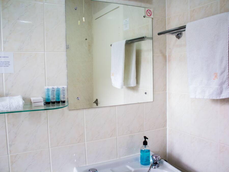 Un baño con un estante de vidrio que sostiene artículos de aseo, un lavamanos con dispensador de jabón y un perchero con una toalla blanca.