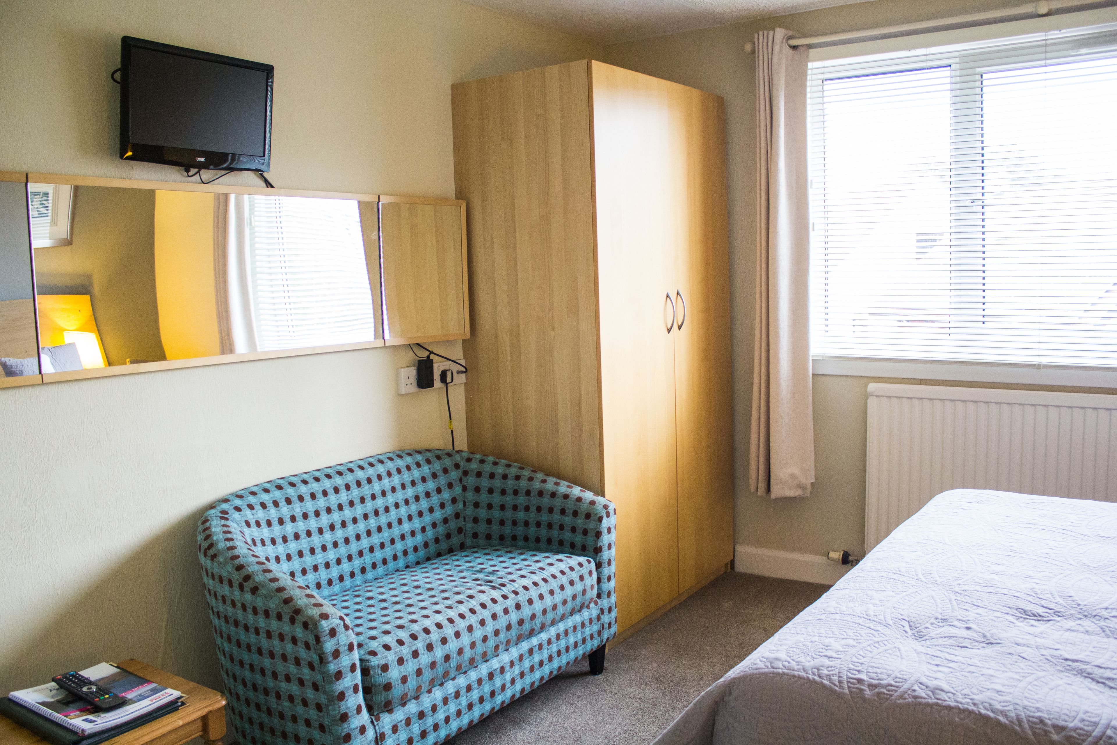 Un dormitorio con cama, una silla azul con lunares, un armario, un televisor en la pared y una ventana con persianas.
