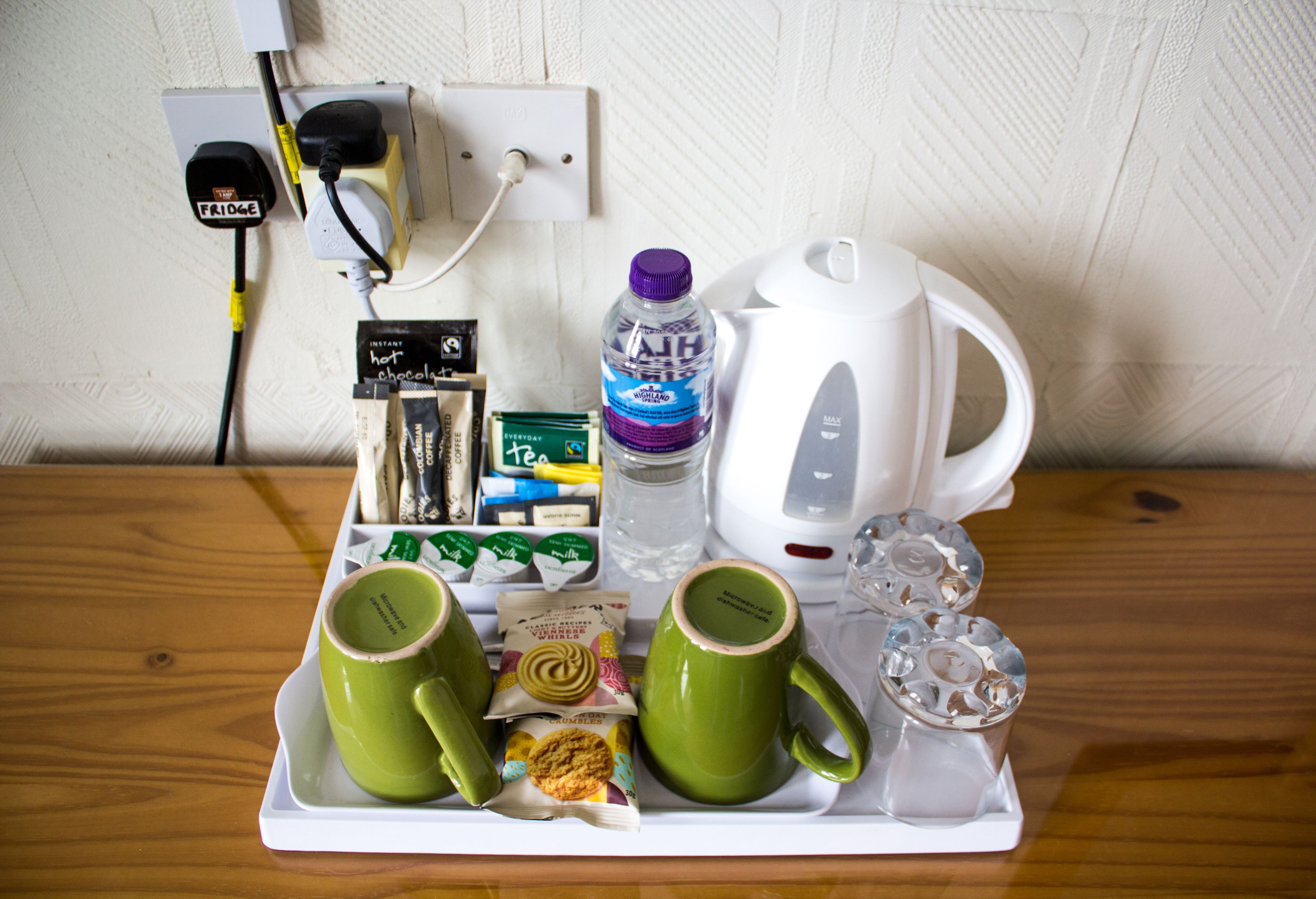 Una bandeja con dos tazas verdes, una botella de agua, una caja de té, un frasco de mermelada y una tetera eléctrica blanca sobre una mesa de madera.