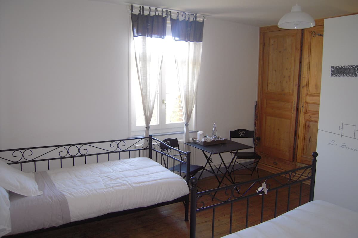 A room with two beds, a small table with a bottle and glasses, a window with curtains, and a wooden door.