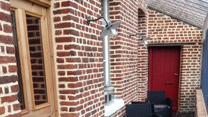 A brick exterior with a wooden door, a red door, and a tiled patio area with wicker furniture. - B&B La Ferme de l'Ostrevent (Estrées)