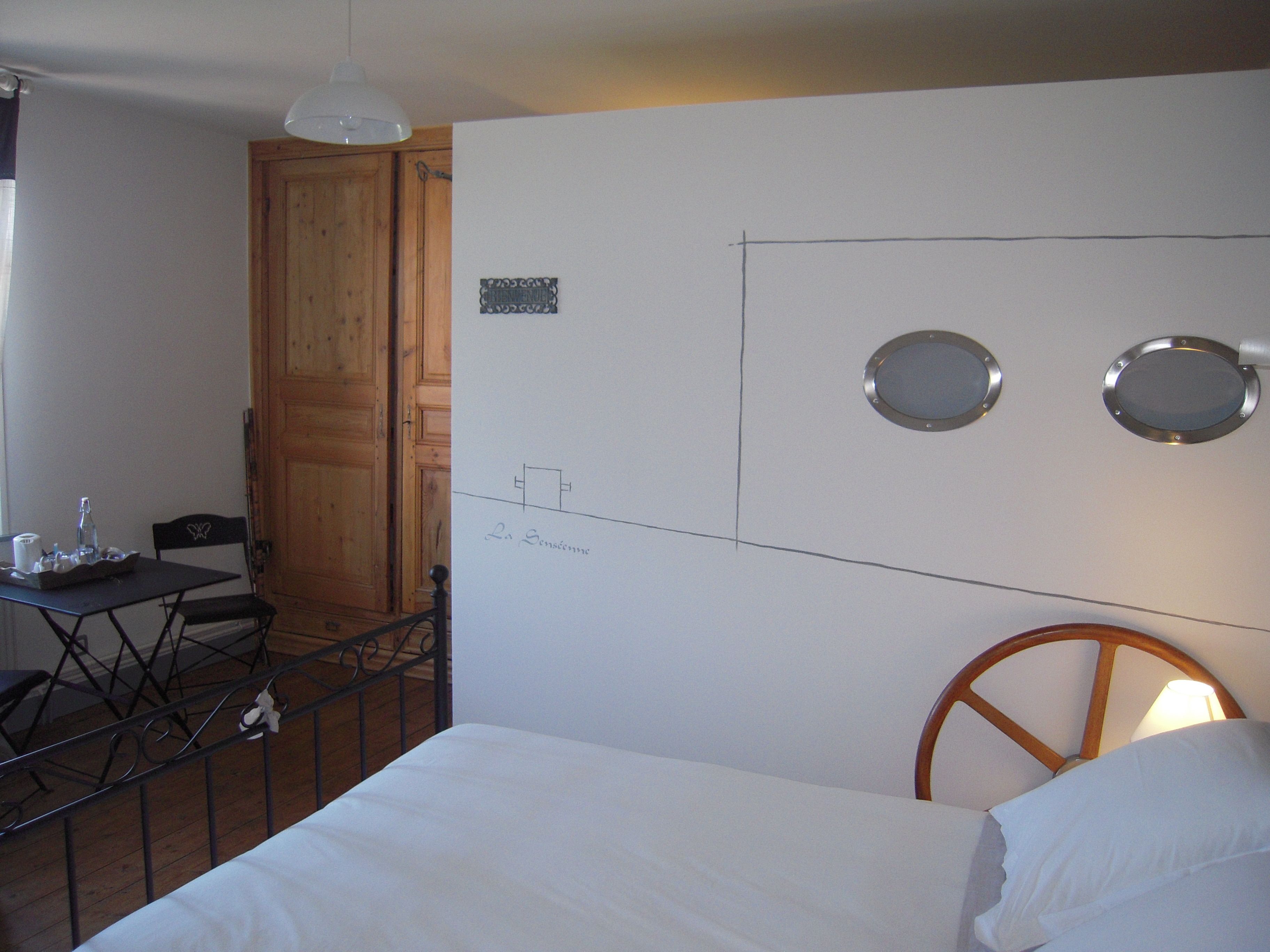 A room with a bed, a desk with a chair, a wooden door, and a wall with a light fixture and two circular mirrors.