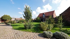 A courtyard with a cobblestone path, a brick building with a red roof, and various shrubs and trees. - B&B La Ferme de l'Ostrevent (Estrées)