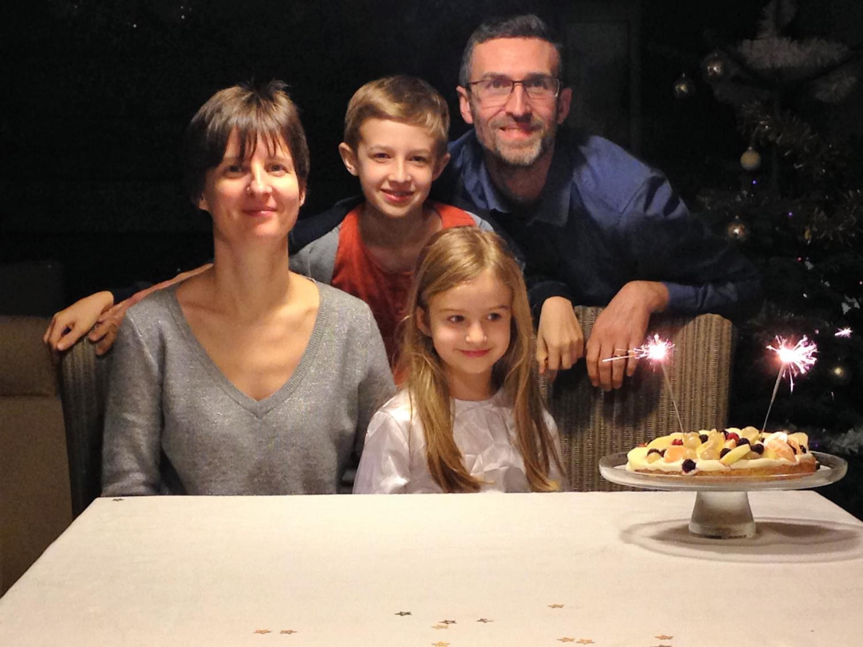 A family seated at a table with a cake featuring sparklers, in front of a Christmas tree.