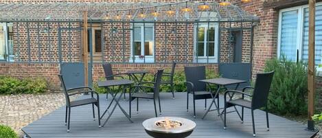 An outdoor patio with a brick building in the background, a pergola with string lights, and outdoor furniture.