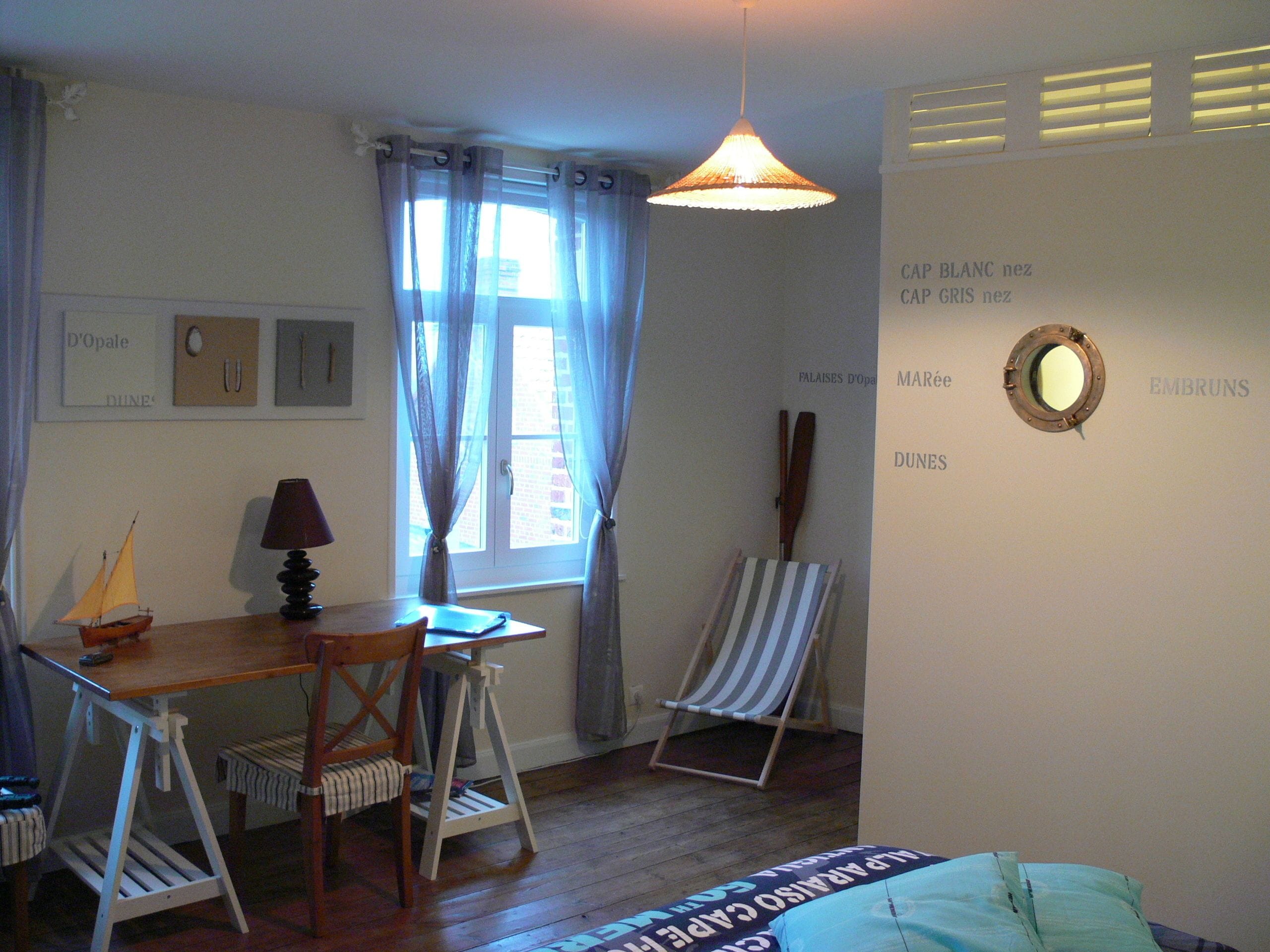 Una habitación con un escritorio de madera, una silla, un pequeño modelo de velero, una lámpara, una ventana con cortinas, una silla de playa a rayas y una pared con texto y un espejo.