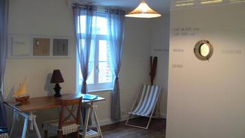 Una habitación con un escritorio de madera, una silla, un pequeño modelo de velero, una lámpara, una ventana con cortinas, una silla de playa a rayas y una pared con texto y un espejo.