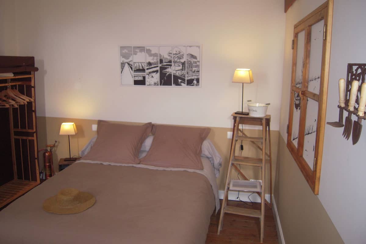 A bedroom with a bed, two bedside lamps, a ladder, a hat on the bed, and a wall-mounted picture.