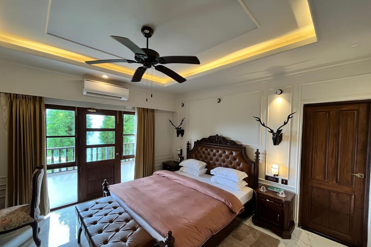 A bedroom with a large bed, a ceiling fan, a wooden door, and a view of greenery outside.