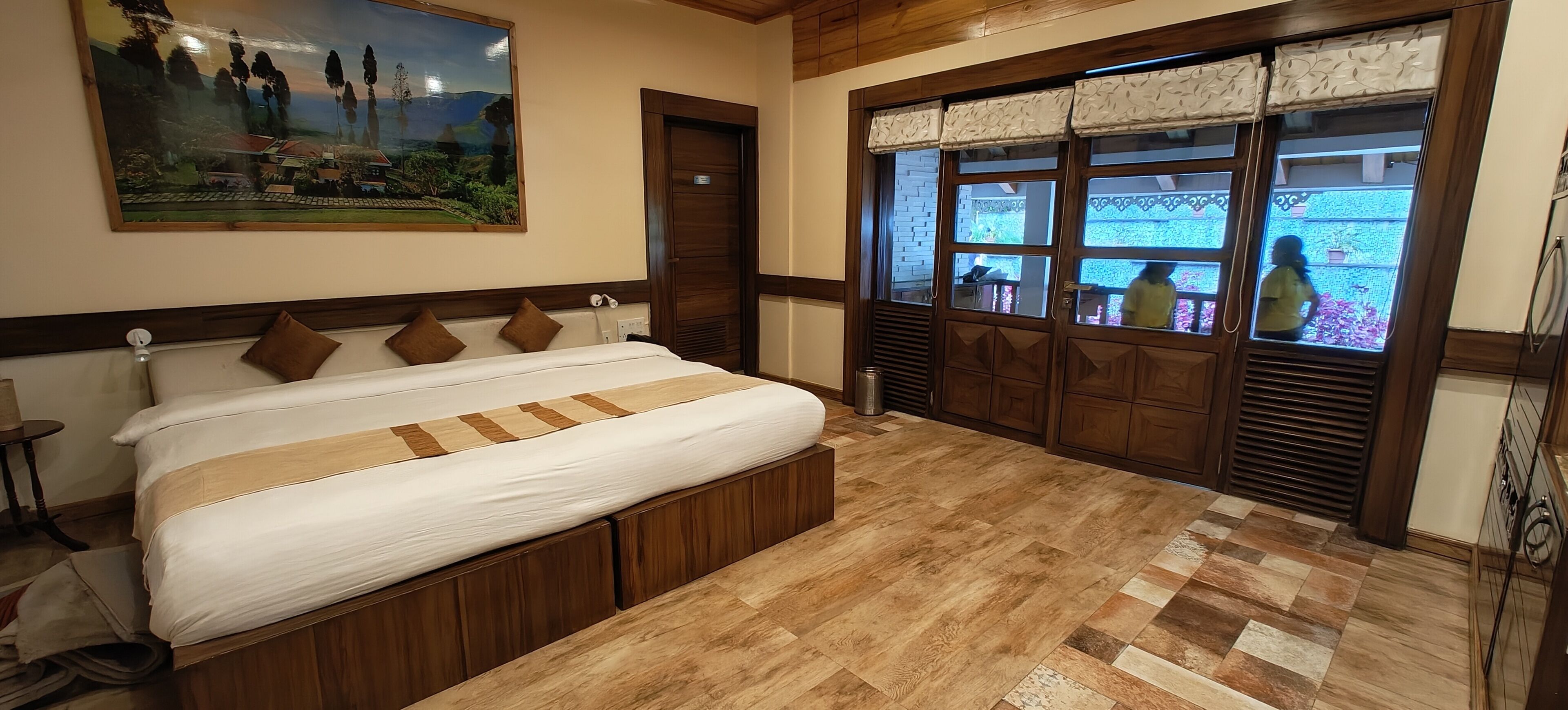 A bedroom with a large bed, wooden flooring, a painting on the wall, and a window with a view of the outside.