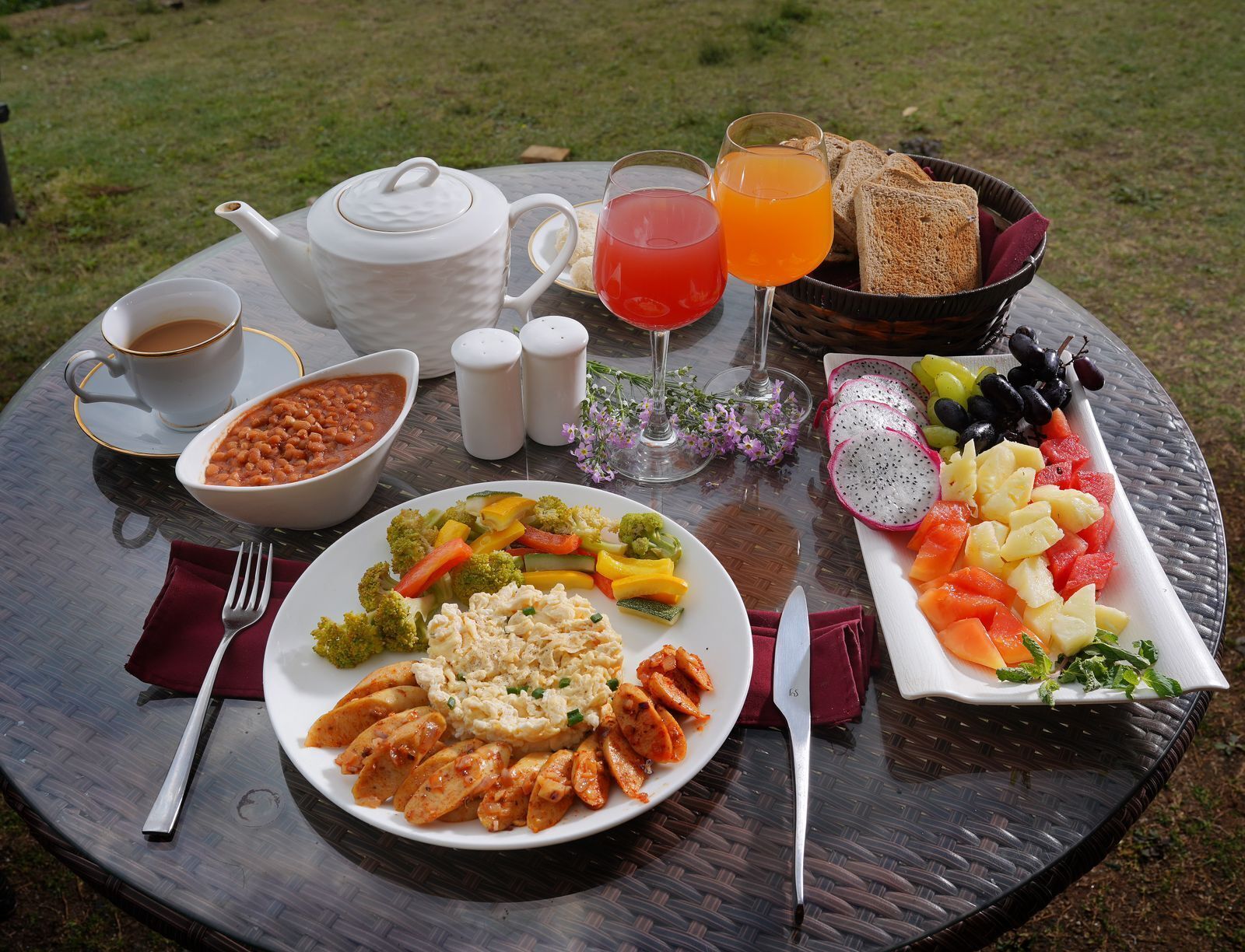 Una mesa redonda de vidrio con una comida que incluye verduras, pollo y una bandeja de frutas, además de bebidas y salsas.