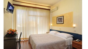 Habitación de hotel con cama, escritorio con computadora, silla, ventana con cortinas y televisor montado en la pared.