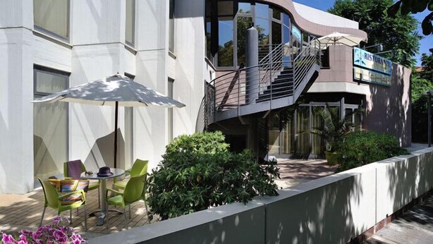 Un edificio moderno con un letrero de restaurante, asientos exteriores con sillas y una sombrilla, y una escalera que lleva a un balcón.