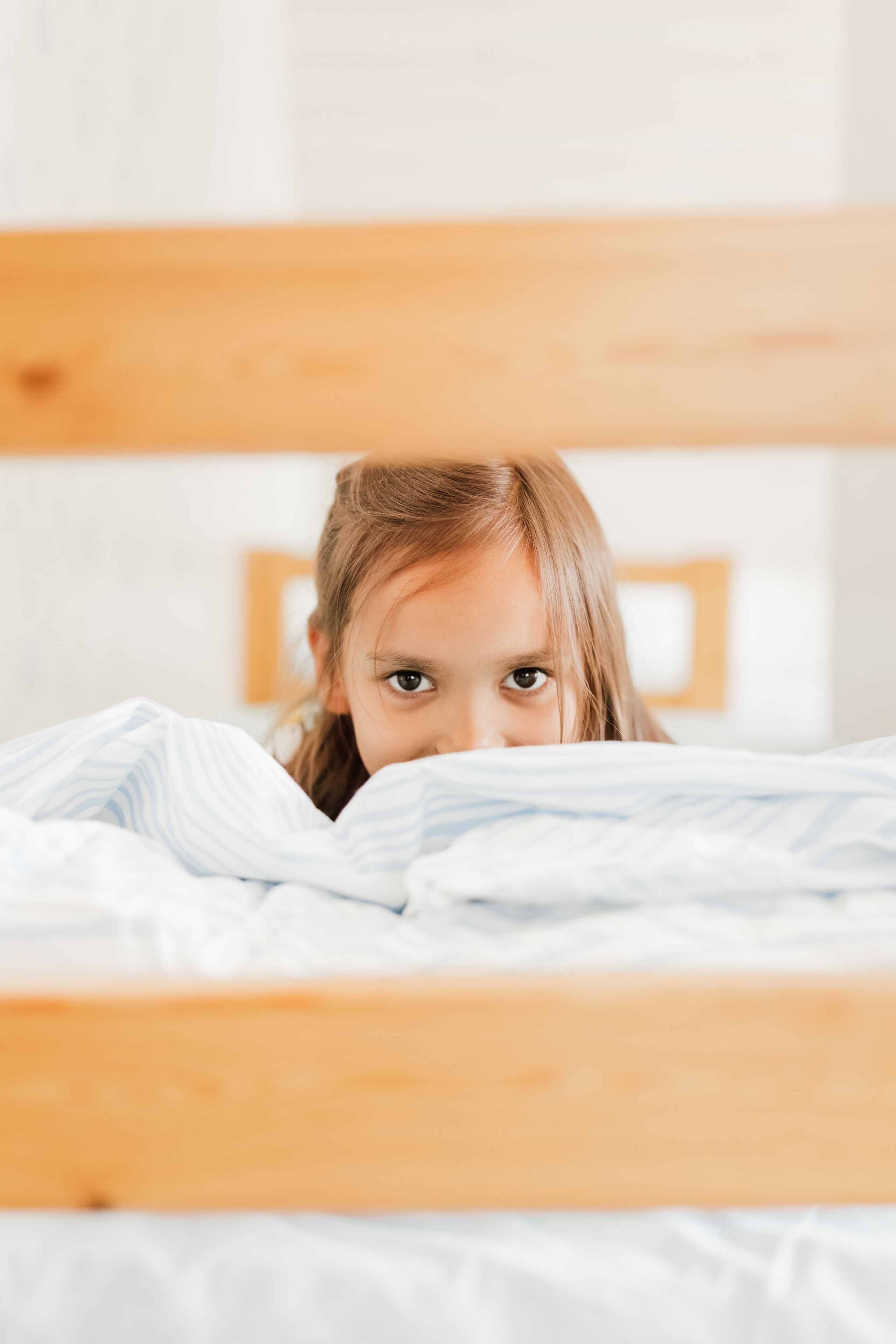 A child peeking over a wooden bed frame with white bedding.