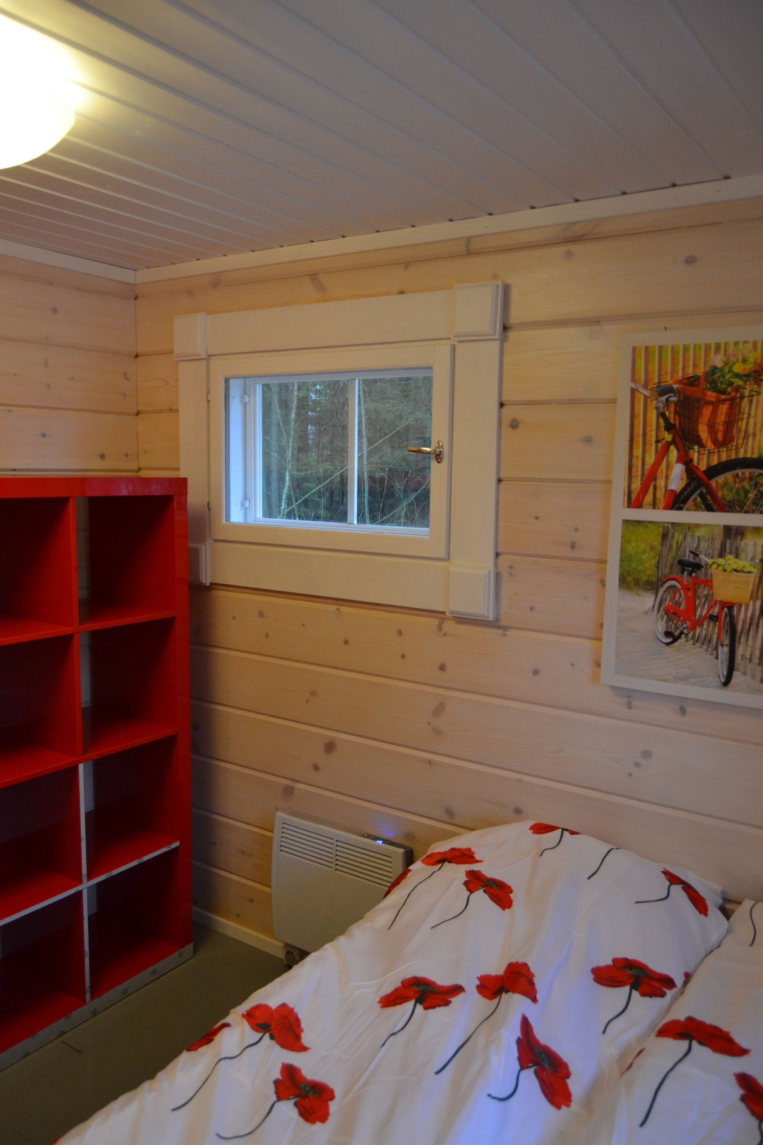 Una stanza tipo baita in legno con una finestra, una scaffalatura rossa e un letto con copripiumino bianco a fiori.