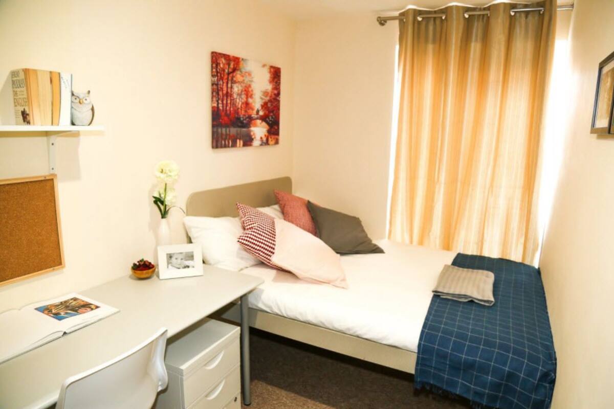 A bedroom with a bed, a desk, a corkboard, a framed picture, and a window with curtains.
