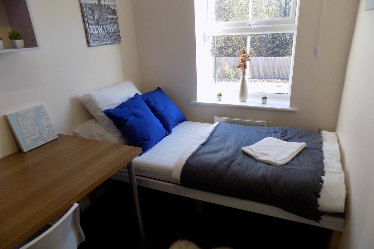 A bedroom with a bed, a desk, a chair, a radiator, and a window with a view of the outside.