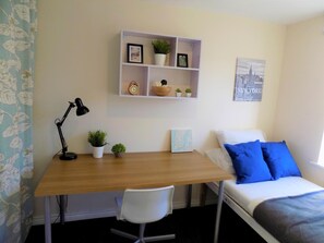 A bedroom with a wooden desk, a white chair, a bed with blue pillows, a wall-mounted shelf with plants and a clock, and a cityscape poster. - Casa Homestay (Coventry)
