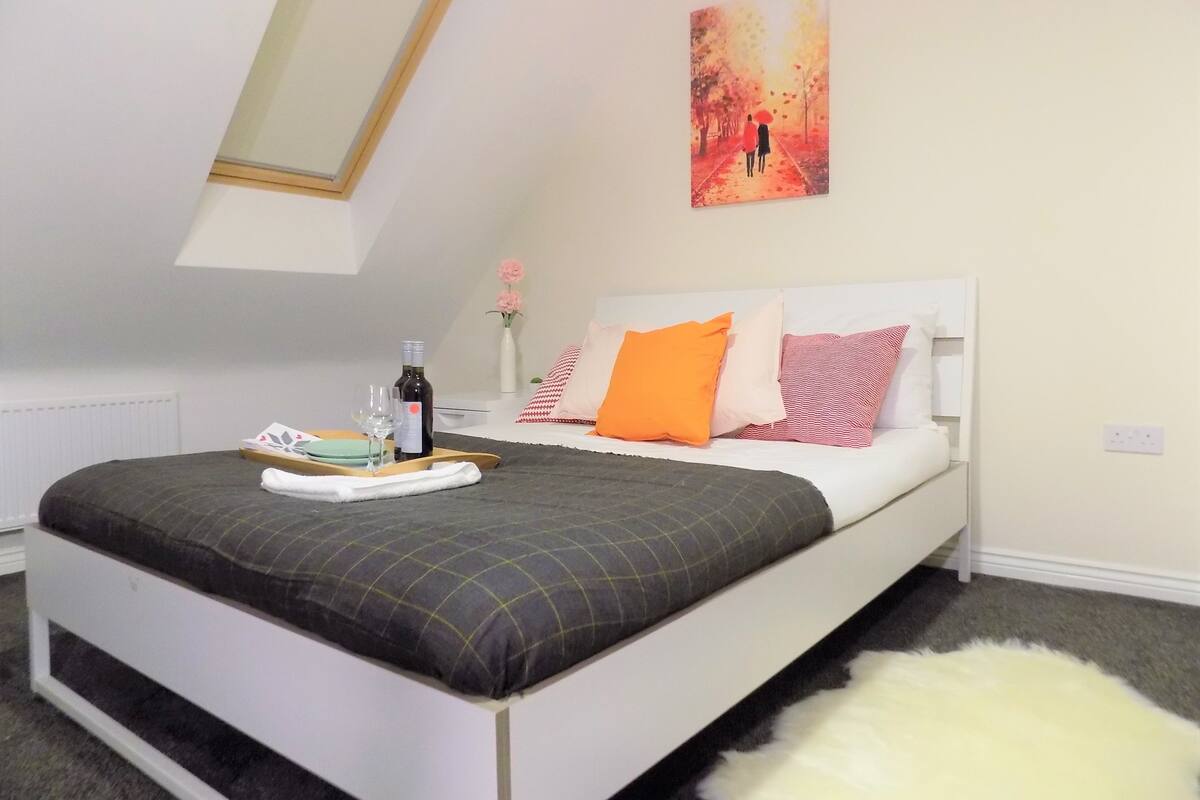 A bedroom with a bed, a bedside table with a bottle and glasses, a painting on the wall, and a small vase with flowers.