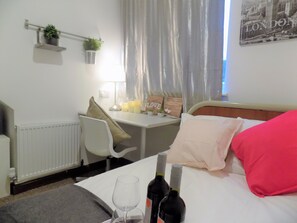 Una camera d'albergo con un letto, una scrivania, una sedia, un termosifone, una finestra con tende, uno scaffale a parete con piante e un quadro appeso al muro.