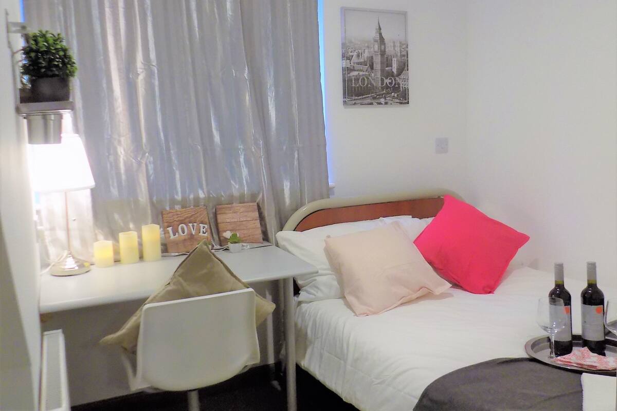 A hotel room with a bed, a desk, a chair, a small table with candles, a potted plant, and a picture of London on the wall.