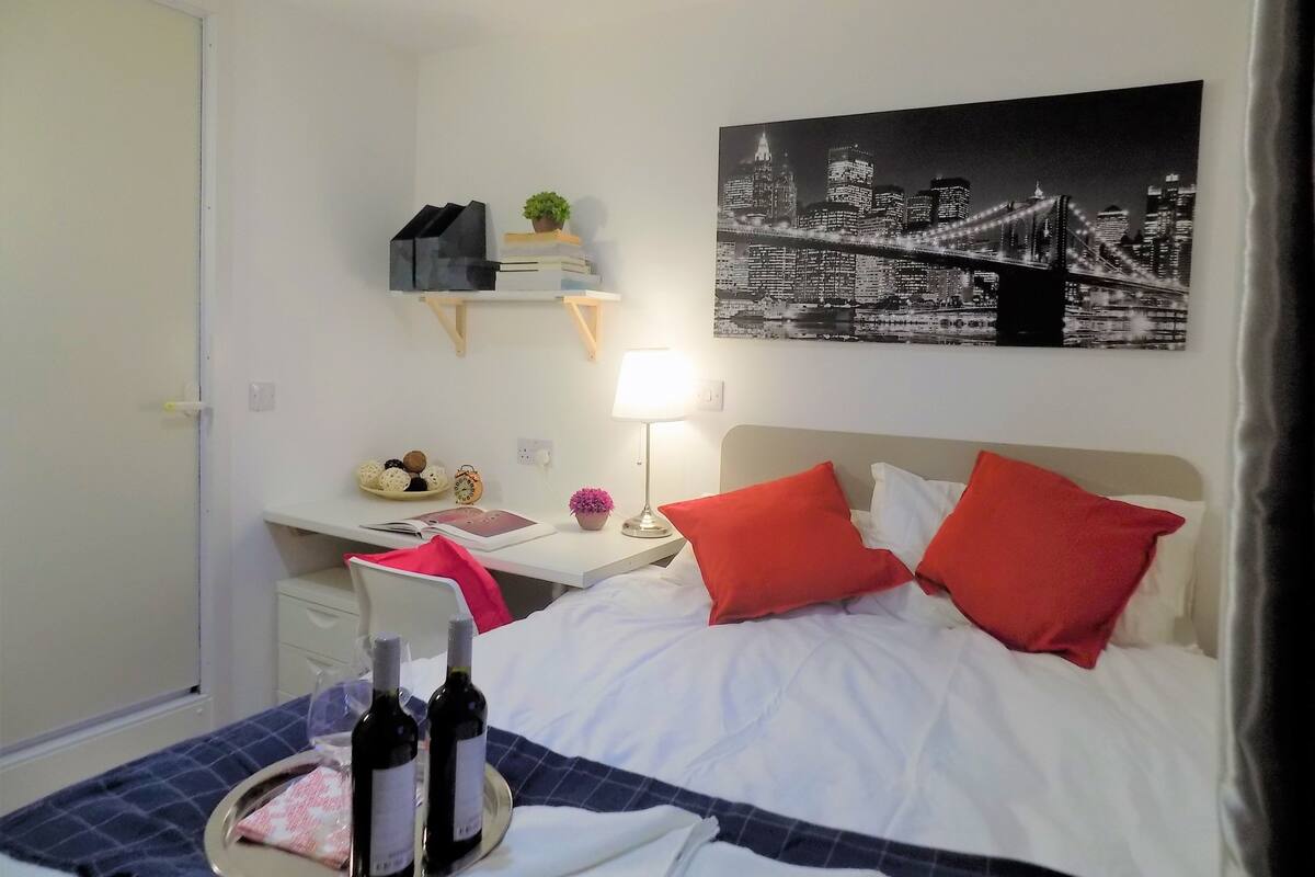 A hotel room with a bed, a desk, a chair, a nightstand, a lamp, a picture of a cityscape, and two bottles of wine on a tray.