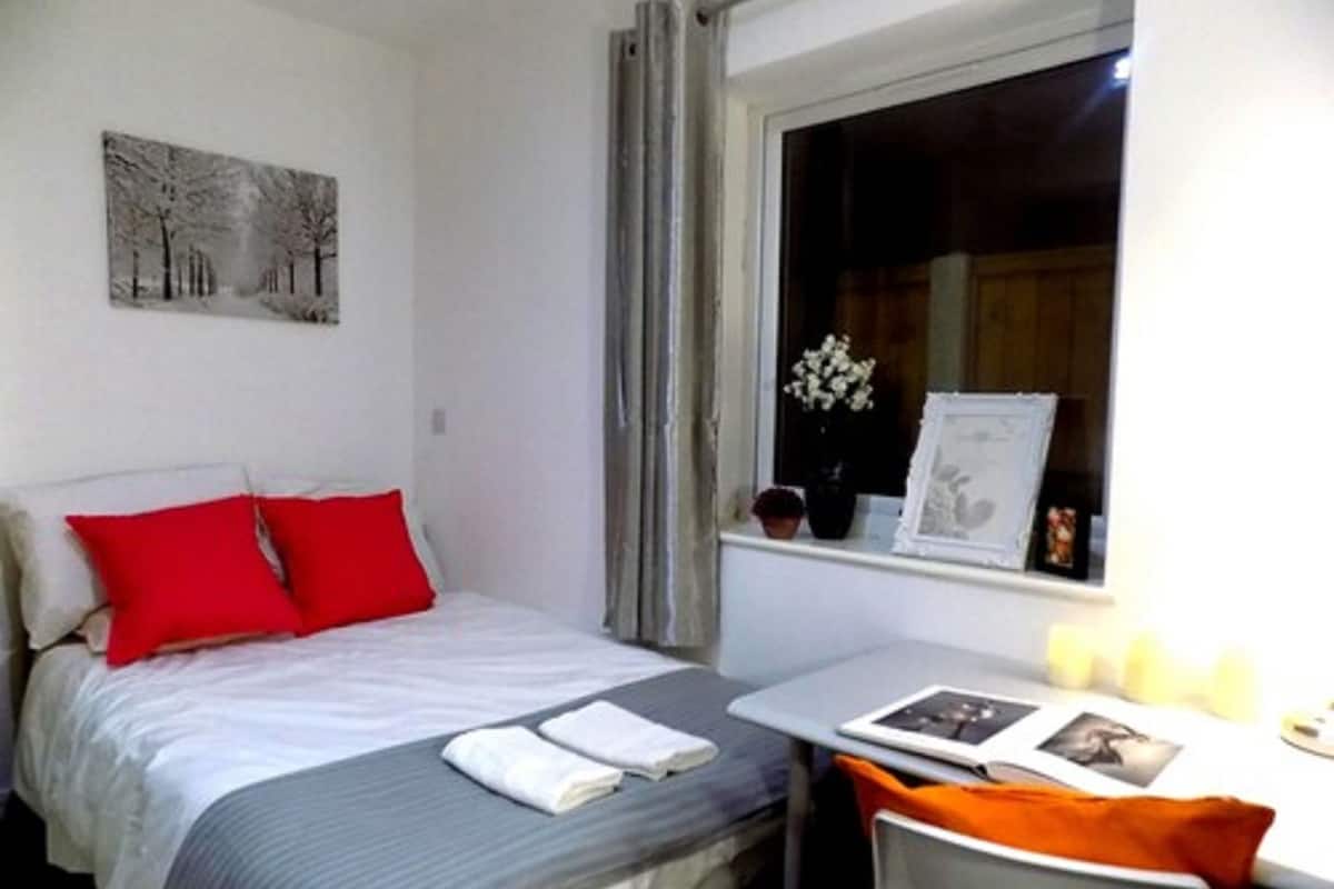 A bedroom with a bed, a bedside table with a lamp, a window with curtains, and a painting on the wall.