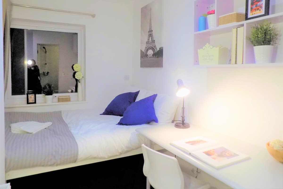 A bedroom with a bed, a desk, a chair, a mirror, and a shelf with books and decorative items.