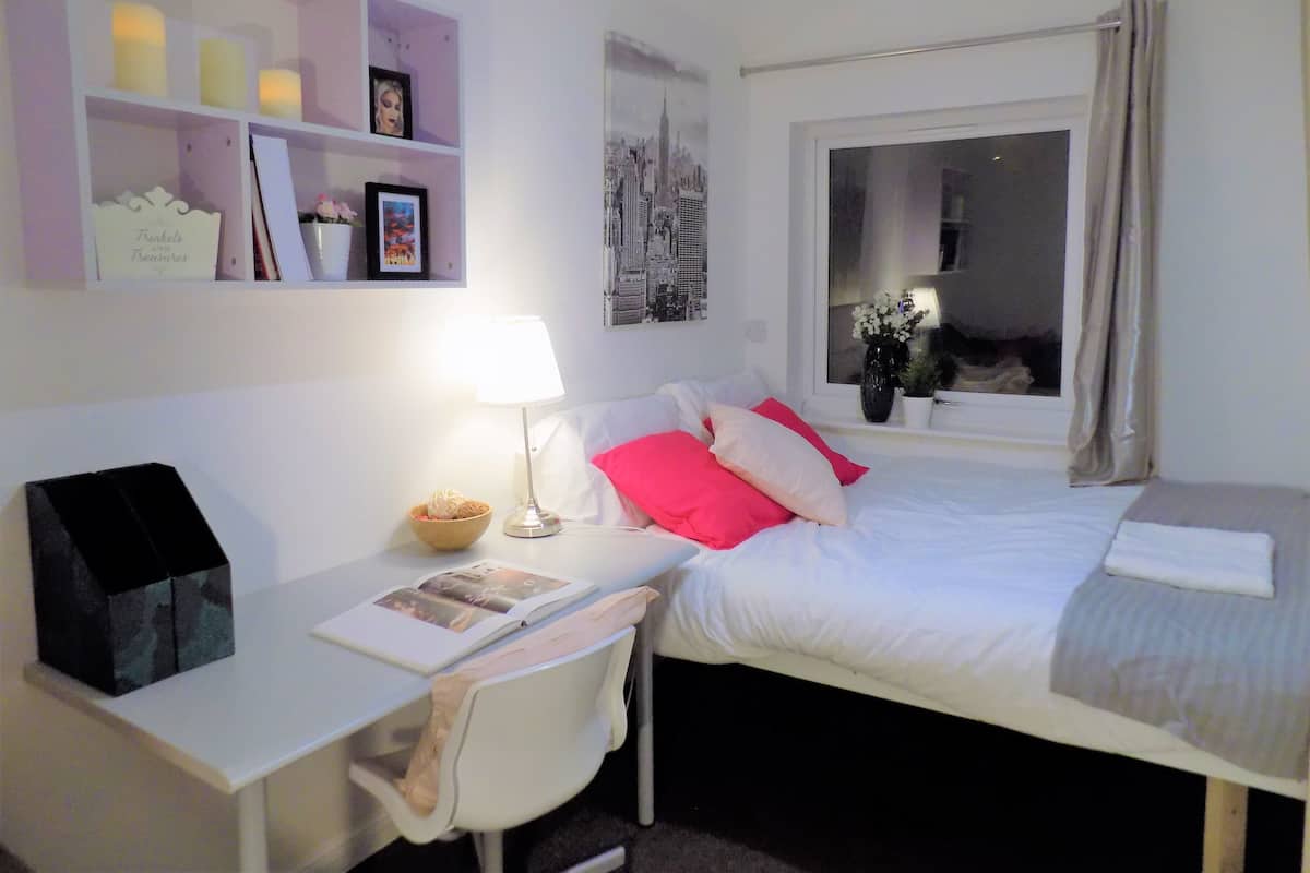 A bedroom with a bed, a desk, a chair, a bedside table with a lamp, a window with curtains, and a wall with framed pictures and a shelf with candles.