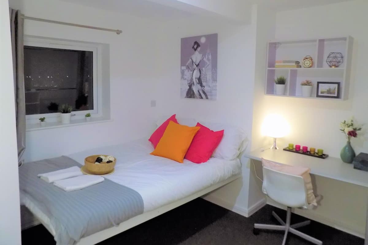 A small, neatly arranged bedroom with a bed, bedside table, lamp, and a window with curtains.